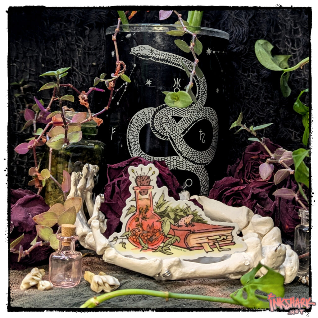 Sticker of a frog hanging out on a spellbook; product is cupped by a skeleton hand in front of dried roses and live plants