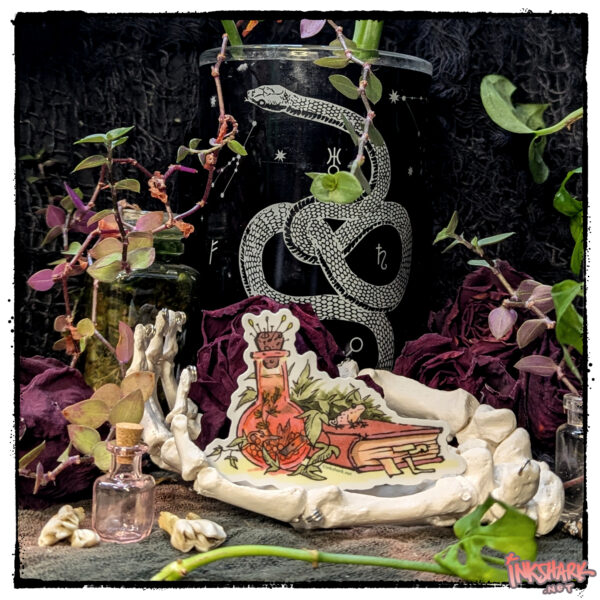 Sticker of a frog hanging out on a spellbook; product is cupped by a skeleton hand in front of dried roses and live plants