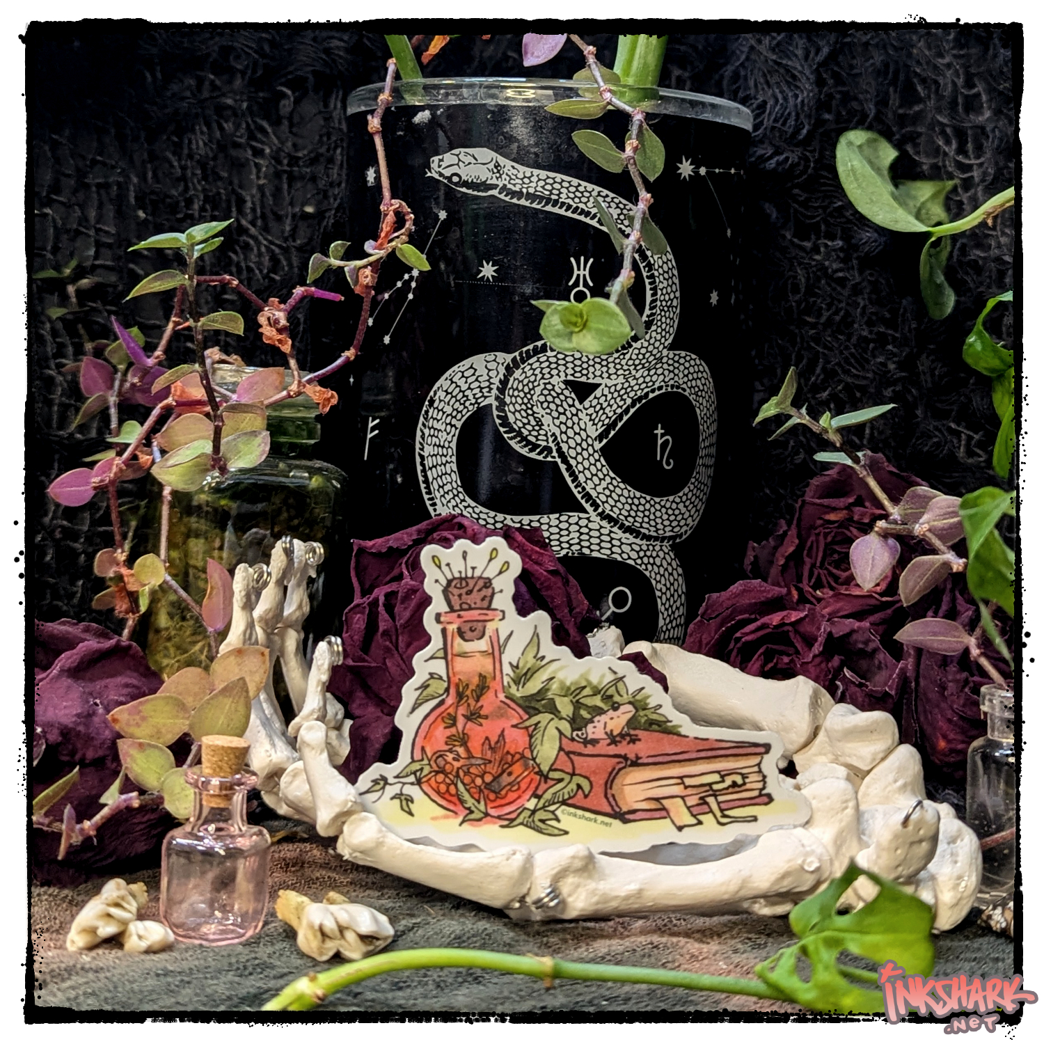 Sticker of a frog hanging out on a spellbook; product is cupped by a skeleton hand in front of dried roses and live plants