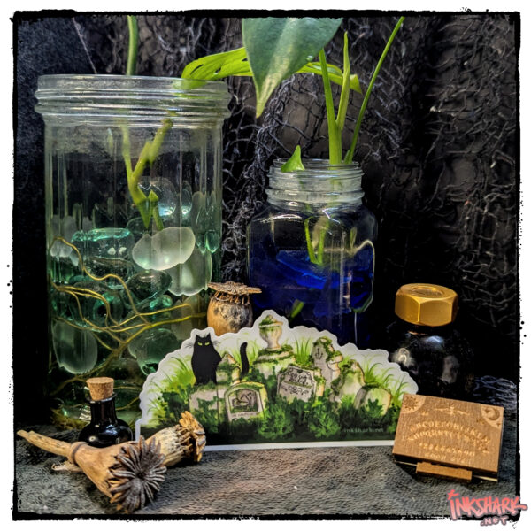 Sticker of a black cat guarding overgrown graves, posed against live plants in glass jars and the tiniest ouija board in the history of the witchy world