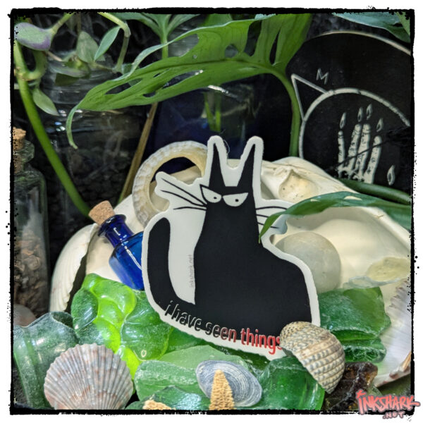 Sticker of a black cat glaring at the viewer; product is perched on a mountain of seaglass, and shells and nestled in foliage
