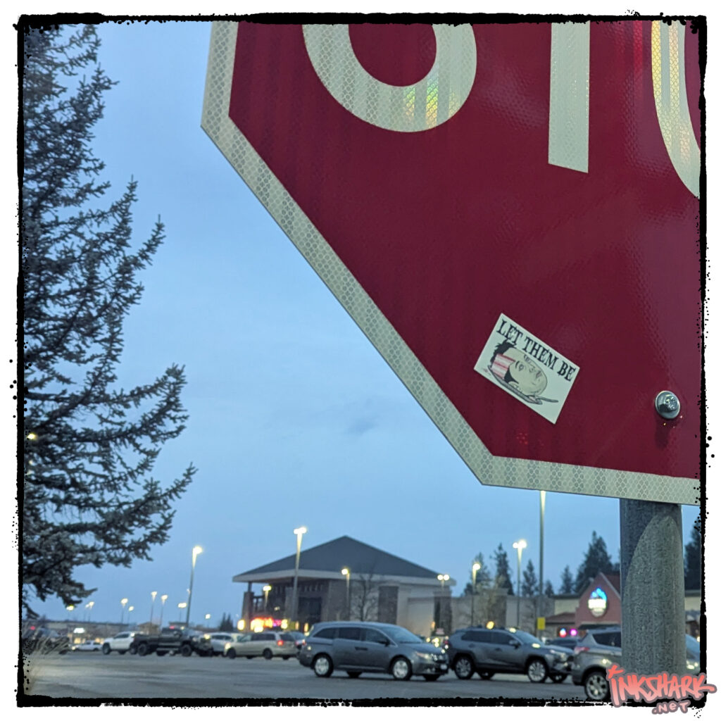 Photo of a sticker on a stop sign. Text says "LET THEM BE" over a drawing of Elon Musk's head with a slice cut out to reveal it's cake