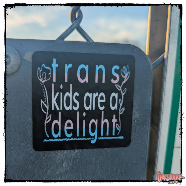 Text colored like the trans Photo of a sticker featuring a flag on a black bg that reads "trans kids are a delight" bookended by scribbly flowers